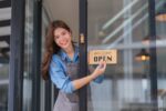 Happy young adult woman small business owner entrepreneur smiling, standing at cafe or restaurant entrance, hanging open sign